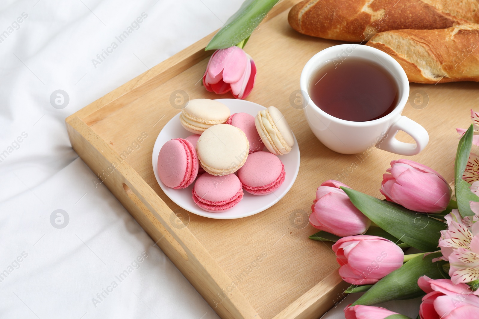 Photo of Tasty breakfast served in bed. Delicious macarons, tea, baguette and flowers on tray