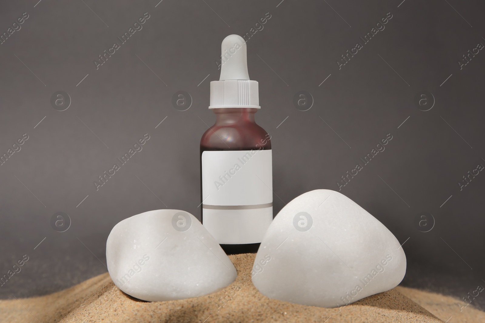 Photo of Bottle of serum and stones on sand against grey background