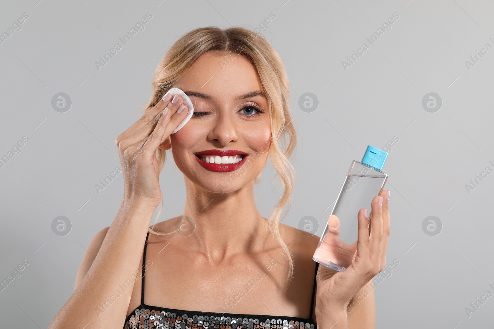 Photo of Smiling woman removing makeup with cotton pad and holding bottle on light grey background