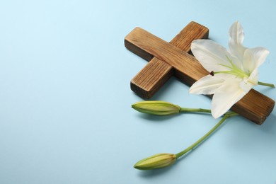 Photo of Wooden cross and lily flowers on light blue background, space for text. Easter attributes