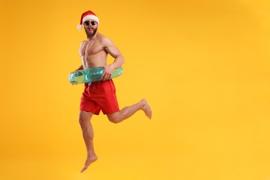 Attractive young man with muscular body in Santa hat with inflatable ring jumping on orange background, space for text