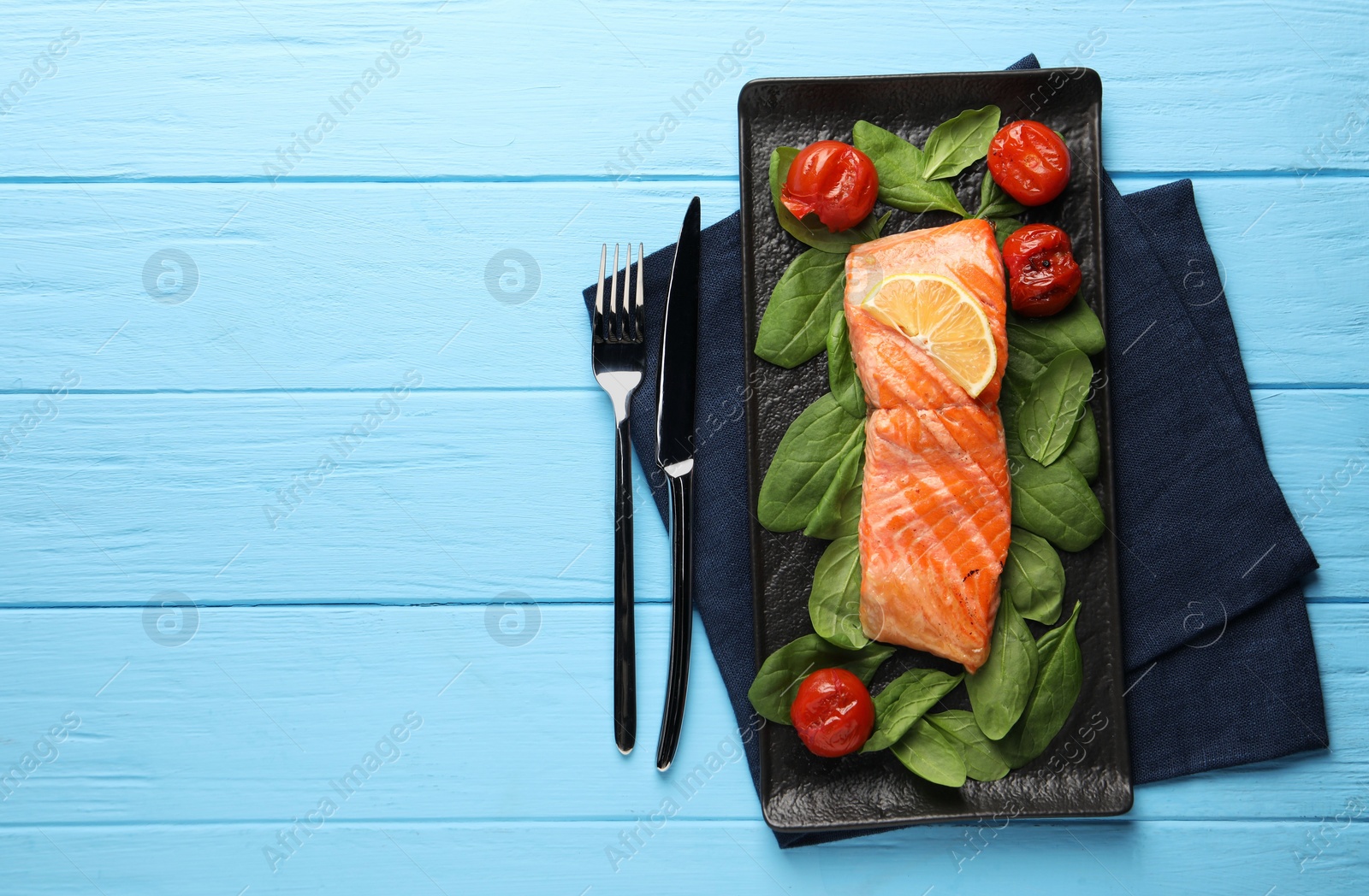 Photo of Tasty grilled salmon with tomatoes, spinach and lemon served on light blue wooden table, top view. Space for text