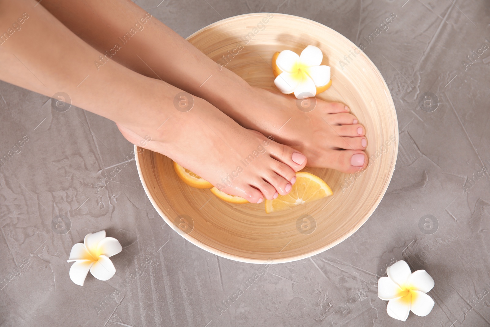 Photo of Woman soaking her feet in bowl with water, orange slices and flower on grey background, top view. Spa treatment