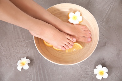 Woman soaking her feet in bowl with water, orange slices and flower on grey background, top view. Spa treatment