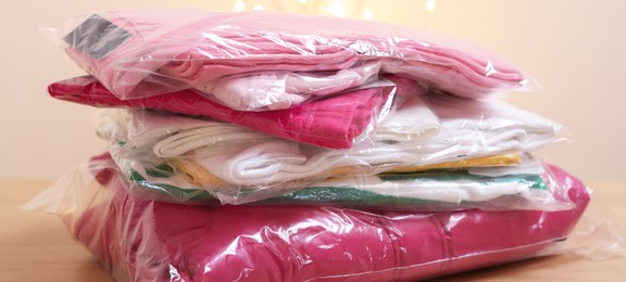 Photo of Stack of folded clothes in plastic bags on wooden table against beige background, closeup