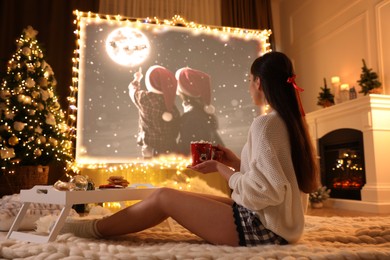 Woman with cup of hot drink watching Christmas movie via video projector in room. Cozy winter holidays atmosphere