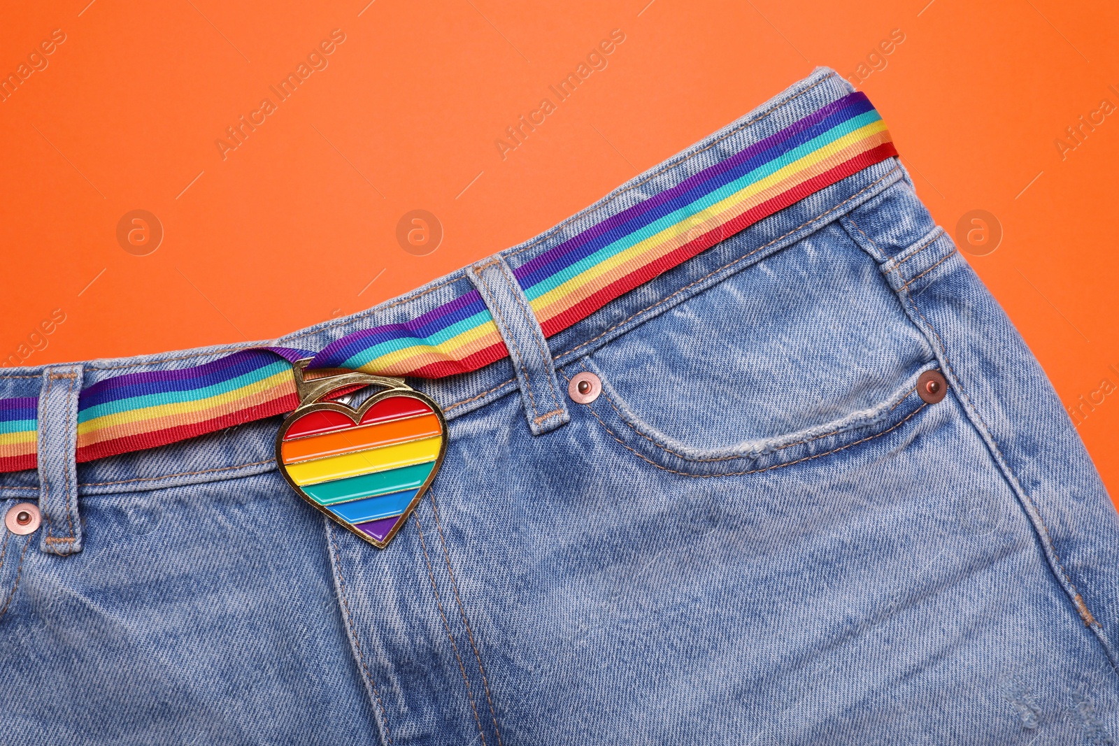 Photo of Jeans and rainbow ribbon with heart shaped pendant on orange background, top view. LGBT pride