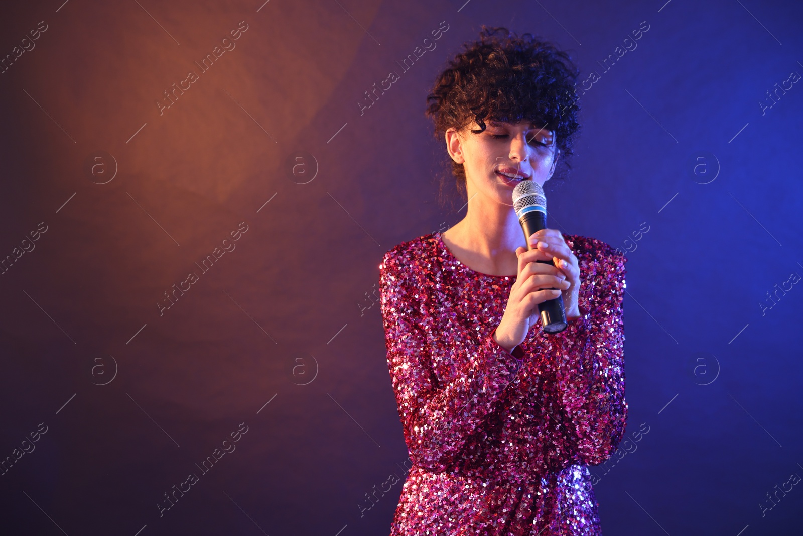 Photo of Beautiful young woman with microphone singing on color background in neon lights. Space for text