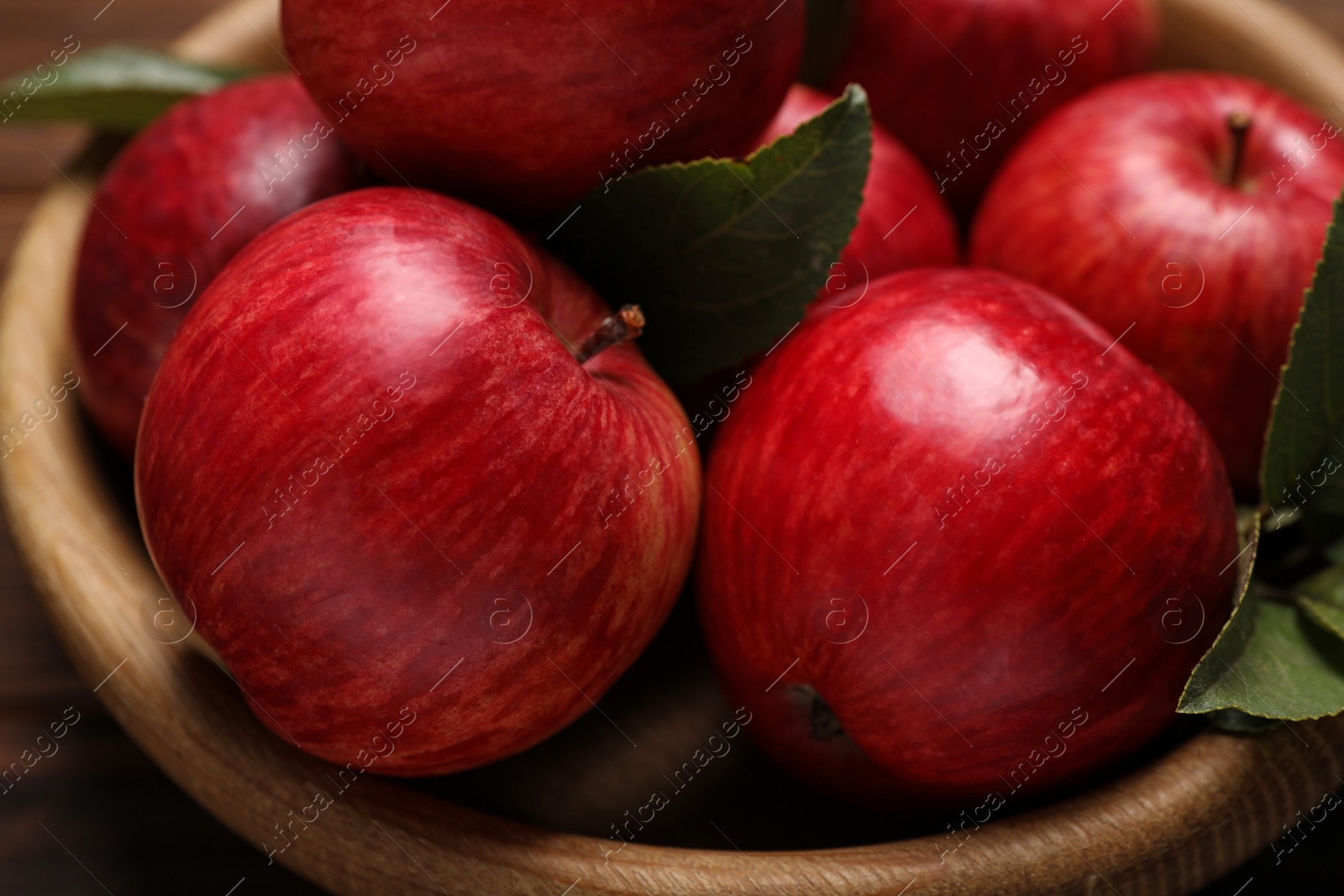 Photo of Ripe red apples and green leaves in wooden bowl on table, closeup