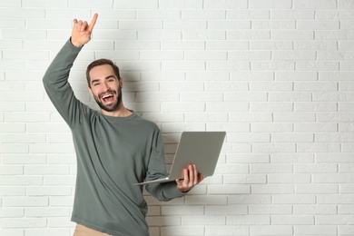 Emotional young man with laptop celebrating victory near brick wall. Space for text