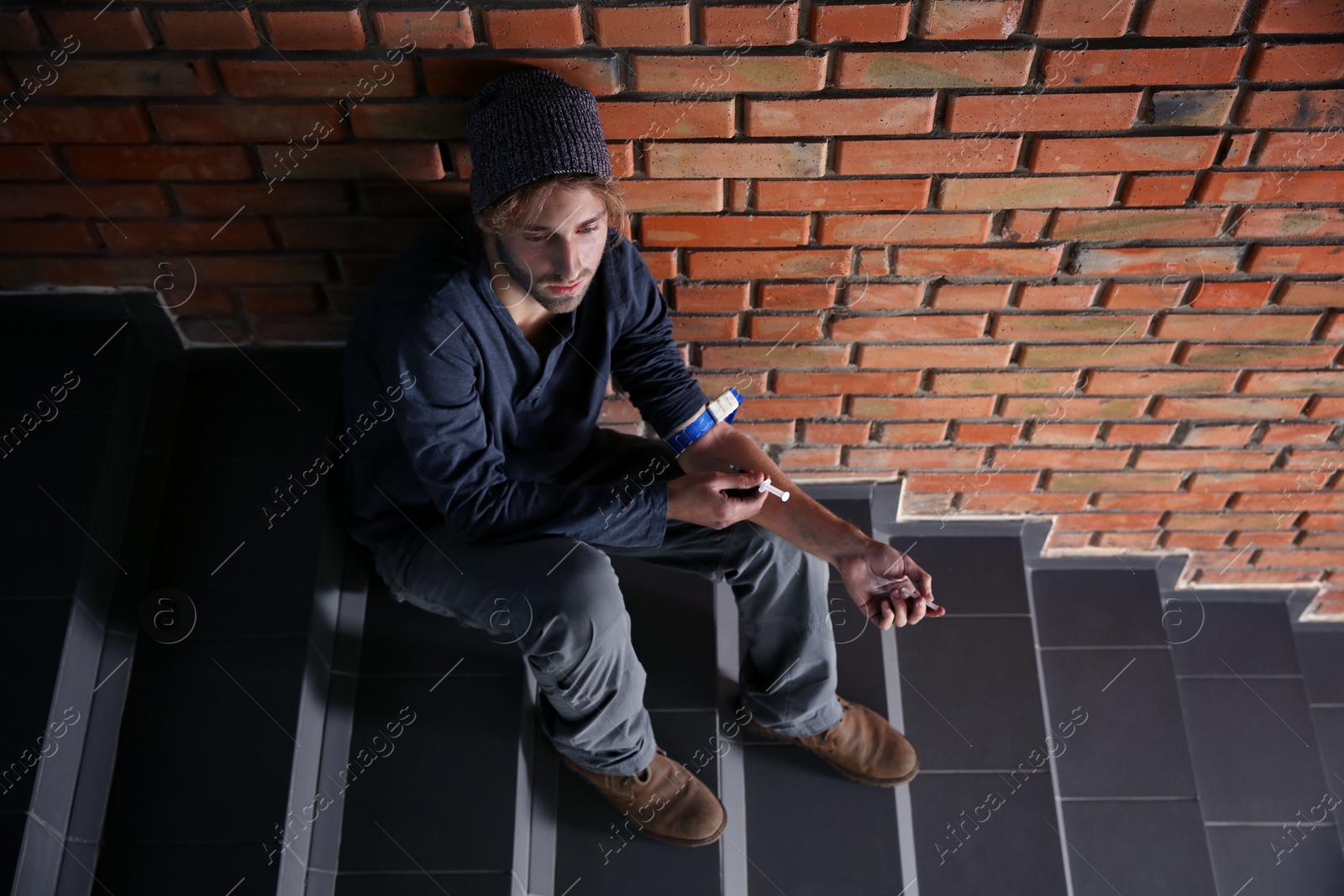 Photo of Male drug addict making injection on stairs near brick wall. Space for text