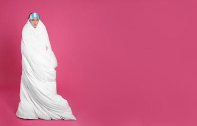 Young woman in sleeping mask wrapped with soft blanket on pink background