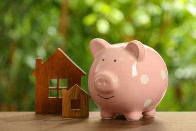 Photo of Piggy bank and house models on wooden table outdoors. Saving money concept