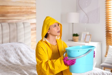 Photo of Young woman in raincoat collecting leaking water from ceiling at home. Time to call roof repair service