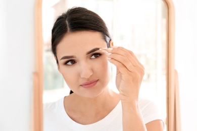 Photo of Young woman plucking eyebrow with tweezers in front of mirror