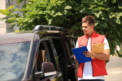 Photo of Young courier with clipboard and envelope near delivery car outdoors