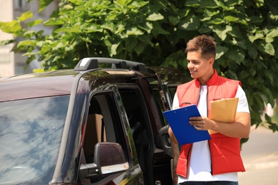 Young courier with clipboard and envelope near delivery car outdoors