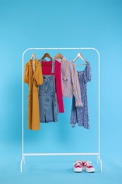 Photo of Rack with stylish women`s clothes on wooden hangers and sneakers against light blue background