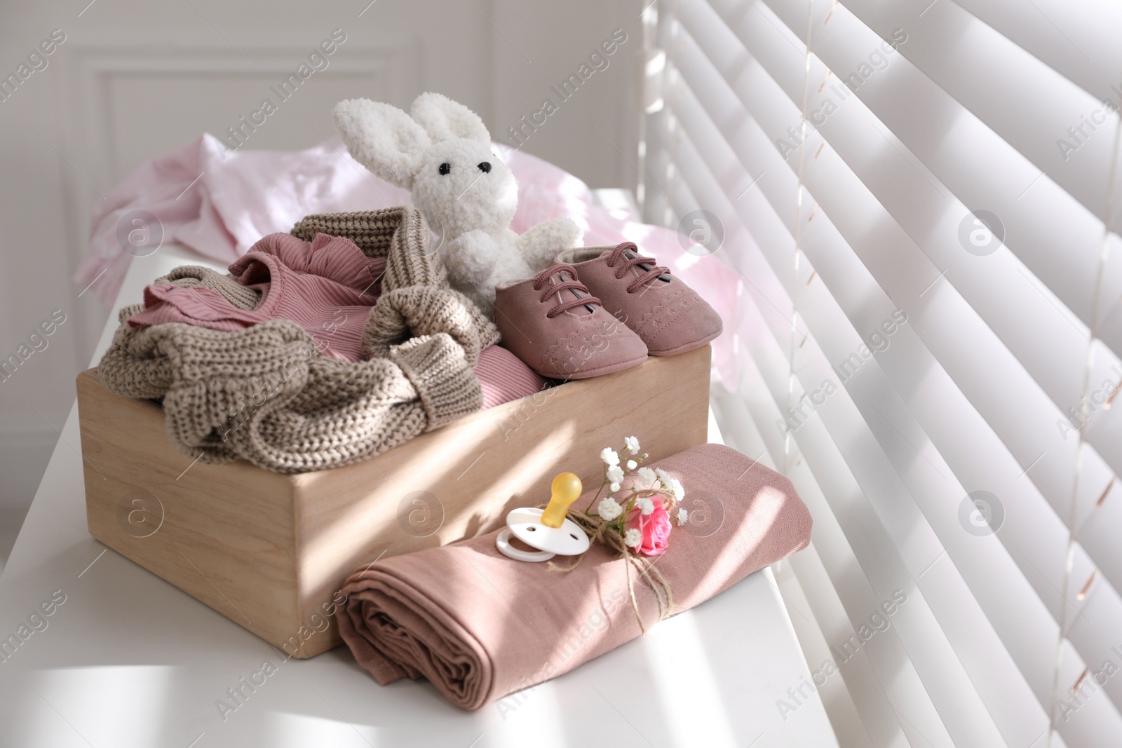 Photo of Wooden crate with children's clothes, shoes, toy and pacifier on white table near window in room
