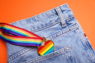 Photo of Jeans and rainbow ribbon with heart shaped pendant on orange background, top view. LGBT pride