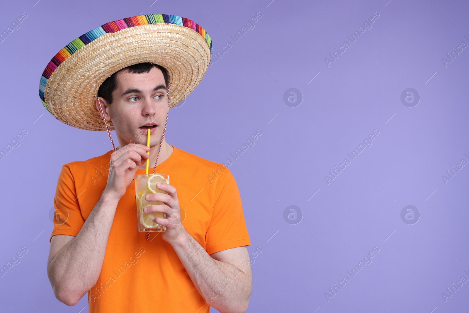 Photo of Young man in Mexican sombrero hat drinking cocktail on violet background. Space for text