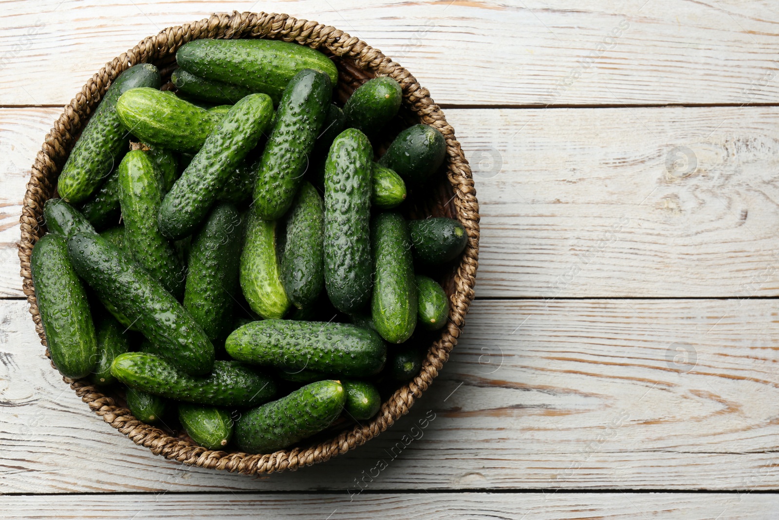 Photo of Fresh ripe cucumbers in wicker basket on white wooden table, top view. Space for text