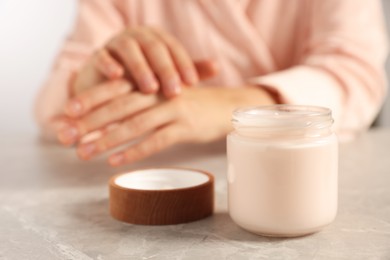 Photo of Woman applying cosmetic cream onto hand at grey table, focus on jar