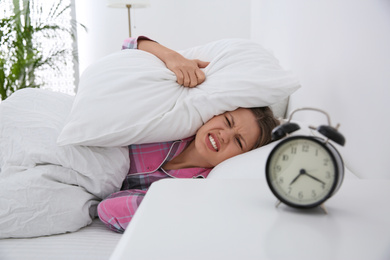 Young woman covering ears with pillows at home in morning, focus on alarm clock