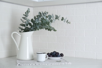 Beautiful eucalyptus branches, cup of drink and fresh plums on countertop in kitchen, space for text. Interior element