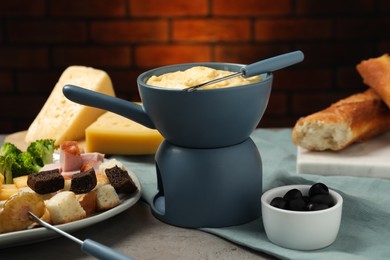 Tasty melted cheese in fondue pot, fork and snacks on grey table, closeup