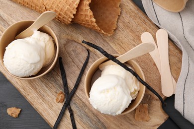 Photo of Paper cups with delicious ice cream and vanilla pods on grey wooden table, flat lay