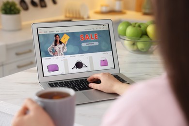 Photo of Woman with cup of tea shopping online during sale on laptop in kitchen, closeup