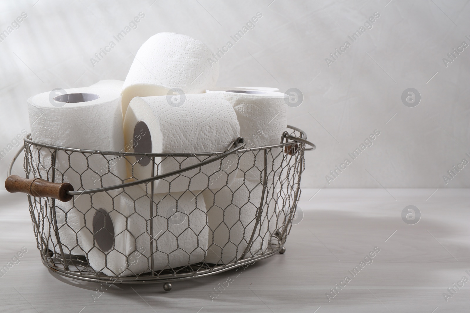 Photo of Soft toilet paper rolls in metal basket on white wooden table, closeup. Space for text