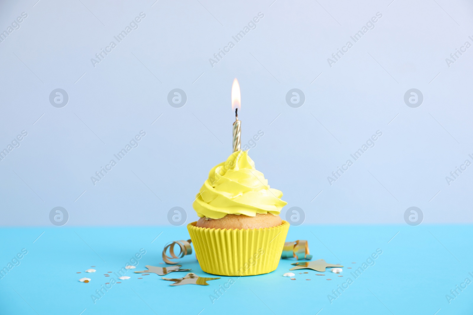Photo of Delicious birthday cupcake with burning candle, streamer and confetti on light blue table against grey background