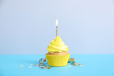 Delicious birthday cupcake with burning candle, streamer and confetti on light blue table against grey background