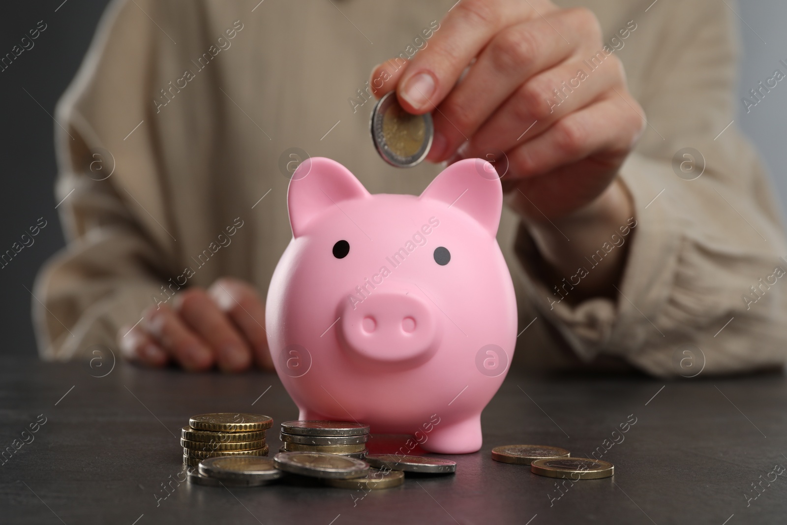 Photo of Woman putting coin into pink piggy bank at black table, closeup