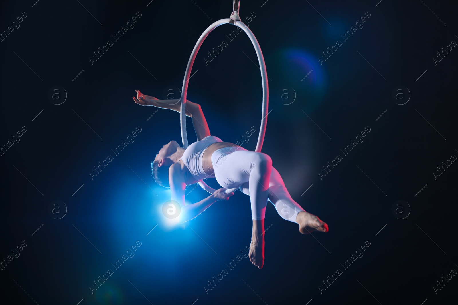 Photo of Young woman performing acrobatic element on aerial ring against dark background