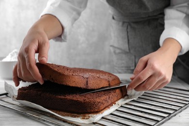 Photo of Woman cutting homemade chocolate cake into layers at white wooden table, closeup