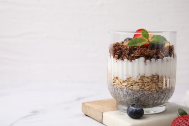 Photo of Tasty granola with berries, yogurt and chia seeds in glass on white marble table, space for text