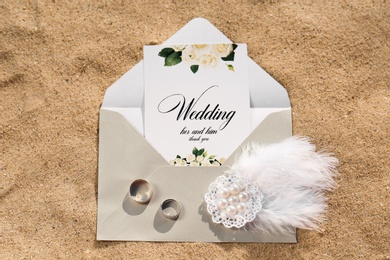 Photo of Envelope with wedding invitation, brooch and gold rings on sandy beach, top view