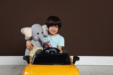 Cute little boy with toy elephant driving children's car near brown wall indoors