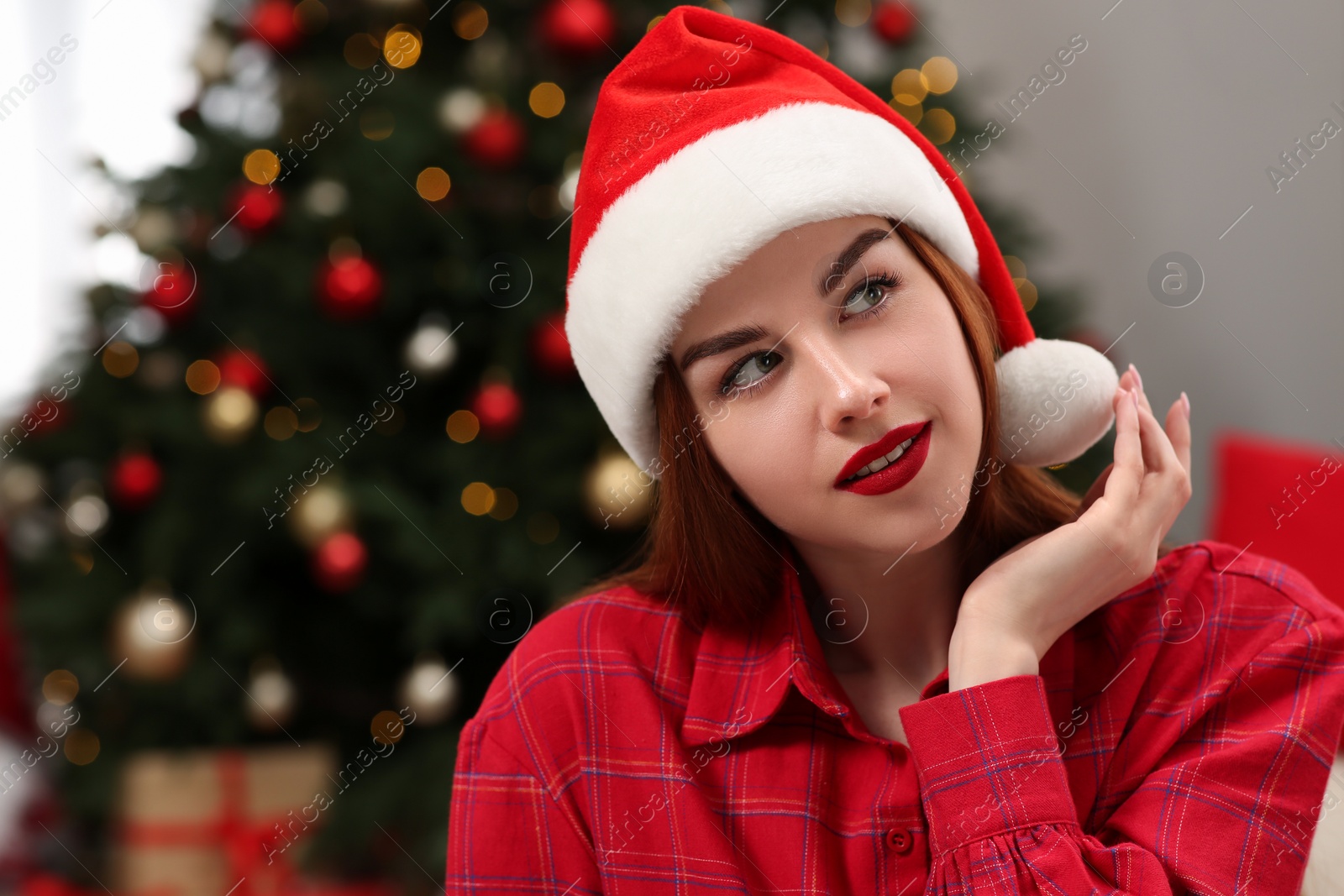 Photo of Beautiful young woman in Santa hat near Christmas tree indoors, space for text