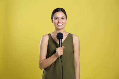 Young female journalist with microphone on yellow background