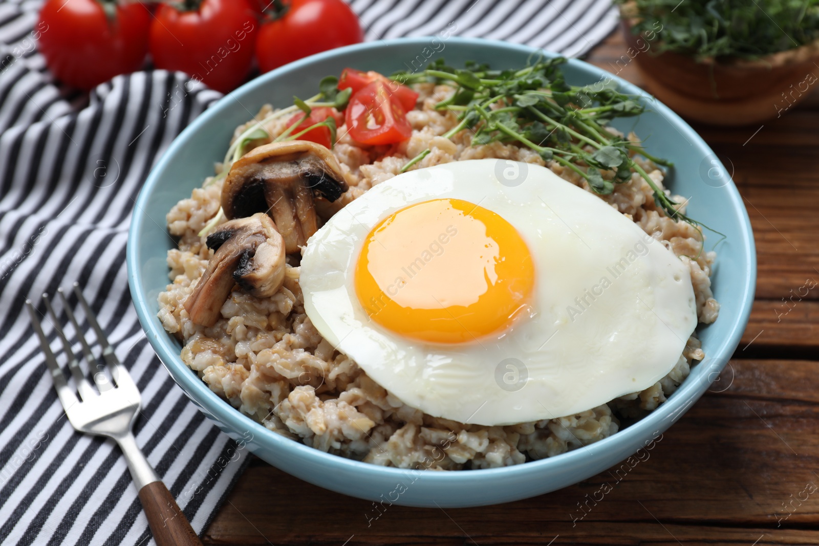 Photo of Tasty boiled oatmeal with fried egg, mushrooms and tomatoes served on wooden table, closeup