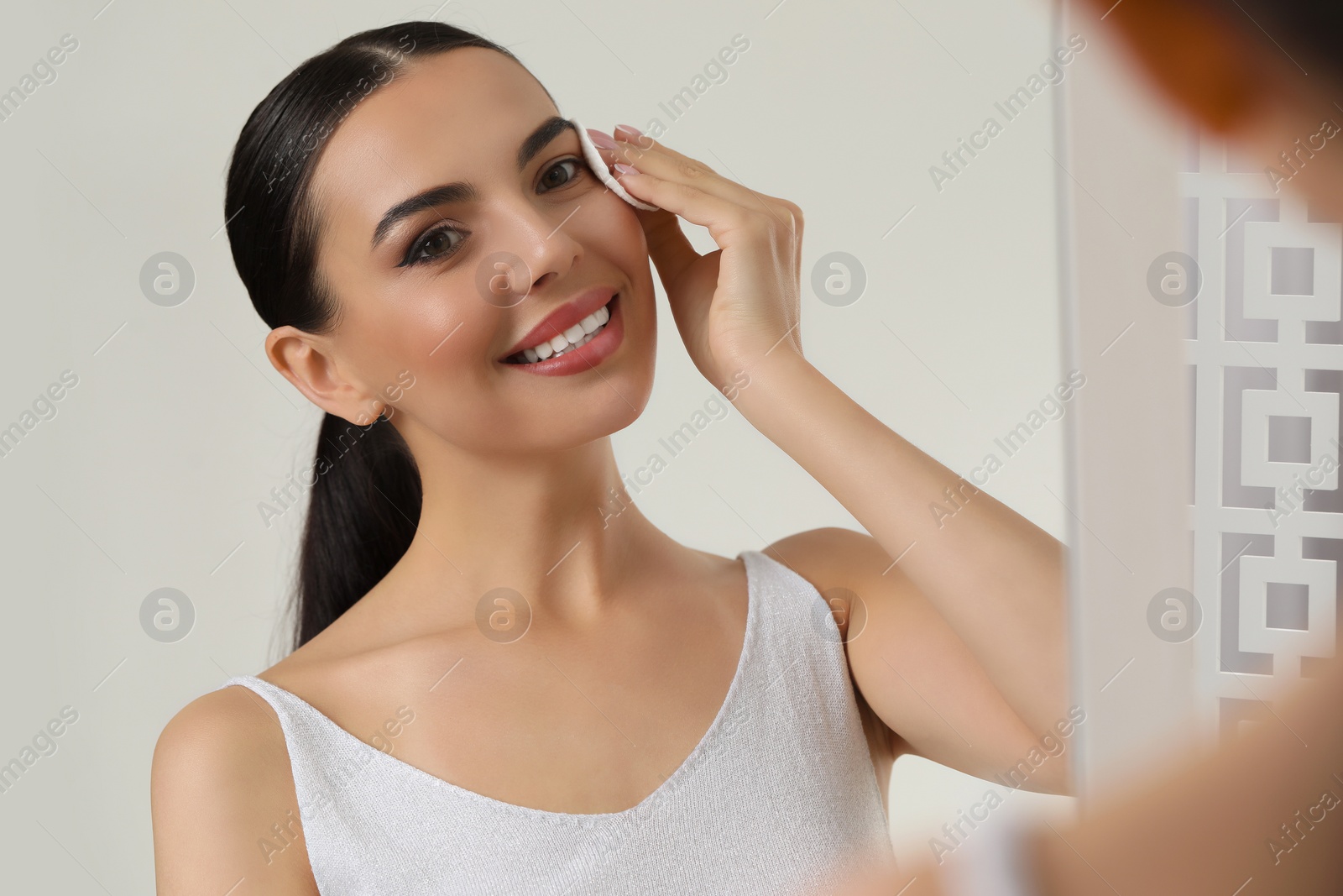 Photo of Beautiful woman removing makeup with cotton pad near mirror indoors