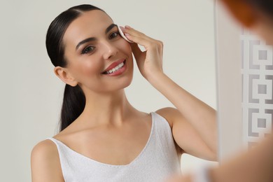 Photo of Beautiful woman removing makeup with cotton pad near mirror indoors