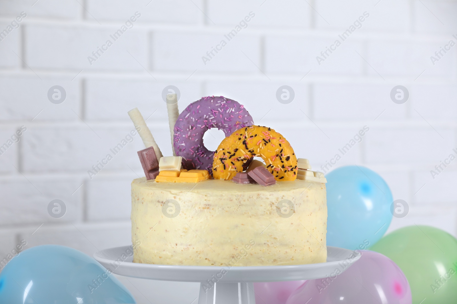 Photo of Delicious cake decorated with sweets and balloons against white brick wall
