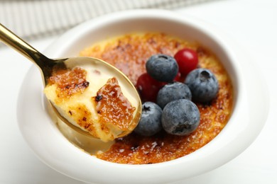 Eating delicious creme brulee with berries from spoon at white table, closeup