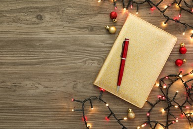 Stylish planner, pen, Christmas lights and balls on wooden background, flat lay with space for text. New Year aims