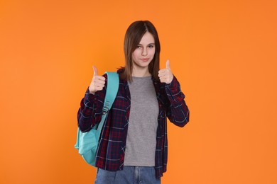 Photo of Portrait of cute teenage girl with backpack showing thumbs up on orange background